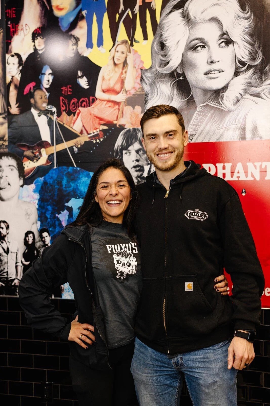 Angela and Chandler Williams smiling in front of their Floyd's 99 Barbershop franchise's collage wall